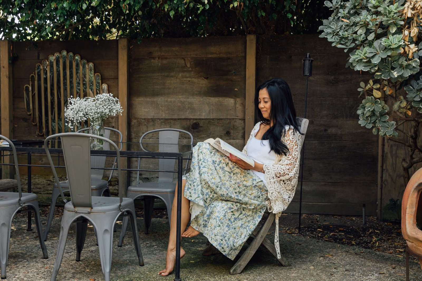 Jaclyn sits reading in a garden chair beside a table of flowers, surrounded by greenery.
