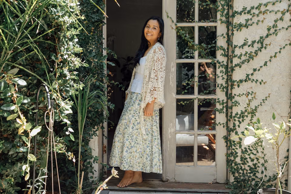 Jaclyn stands barefoot in a sunlit doorway framed by climbing greenery, looking over her shoulder and smiling.