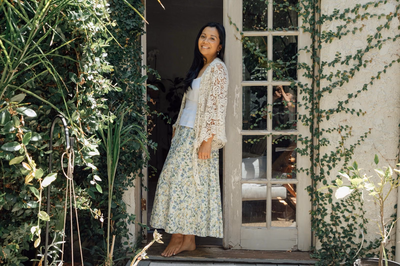 Jaclyn stands barefoot in a sunlit doorway framed by climbing greenery, looking over her shoulder and smiling.