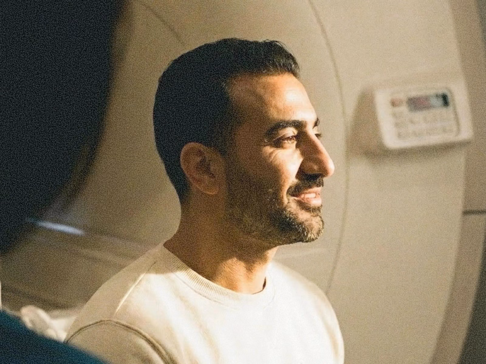 Smiling man in profile facing right, with short dark hair and a trimmed beard, wearing a white crewneck sweater and lit by warm sunlight inside what looks like an MRI scanner room.